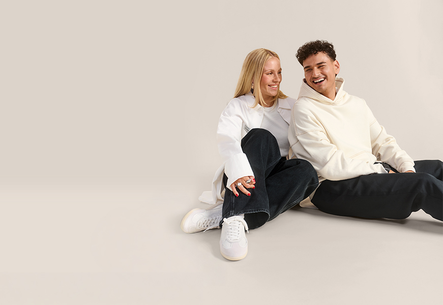 Zwei junge Menschen sitzen auf dem Boden vor einem neutralen, beigen Hintergrund und lachen gemeinsam.